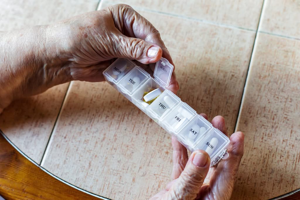 AdobeStock_1919591586 Close up senior woman hands holding weekly pill organizer with tablets sorted by days
